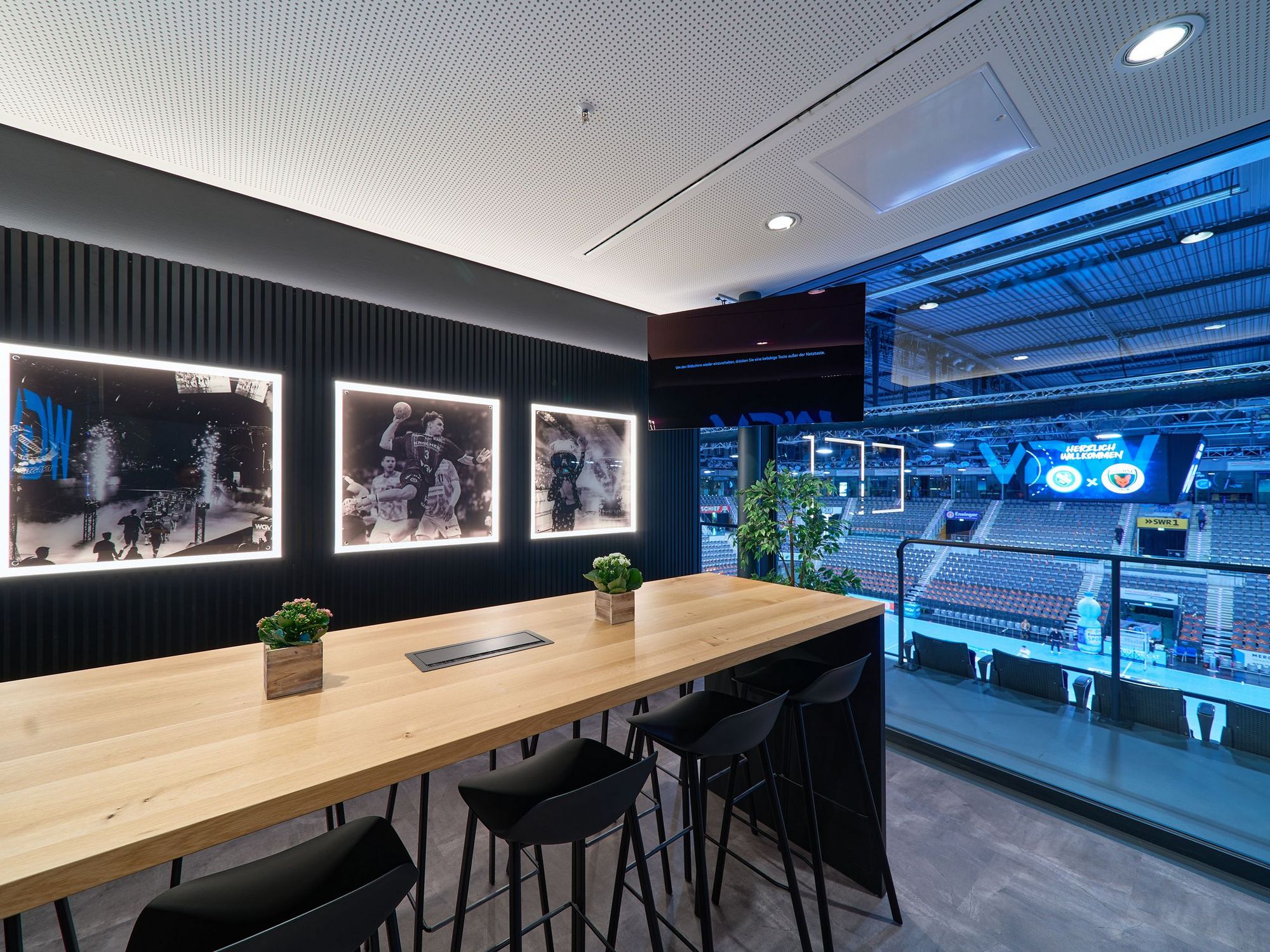Eine Loge mit langem Holztisch und Barhockern. An der Wand hängen Bilder vom Handball. Die große Glasfront ermöglicht den direkten Blick in die Porsche-Arena