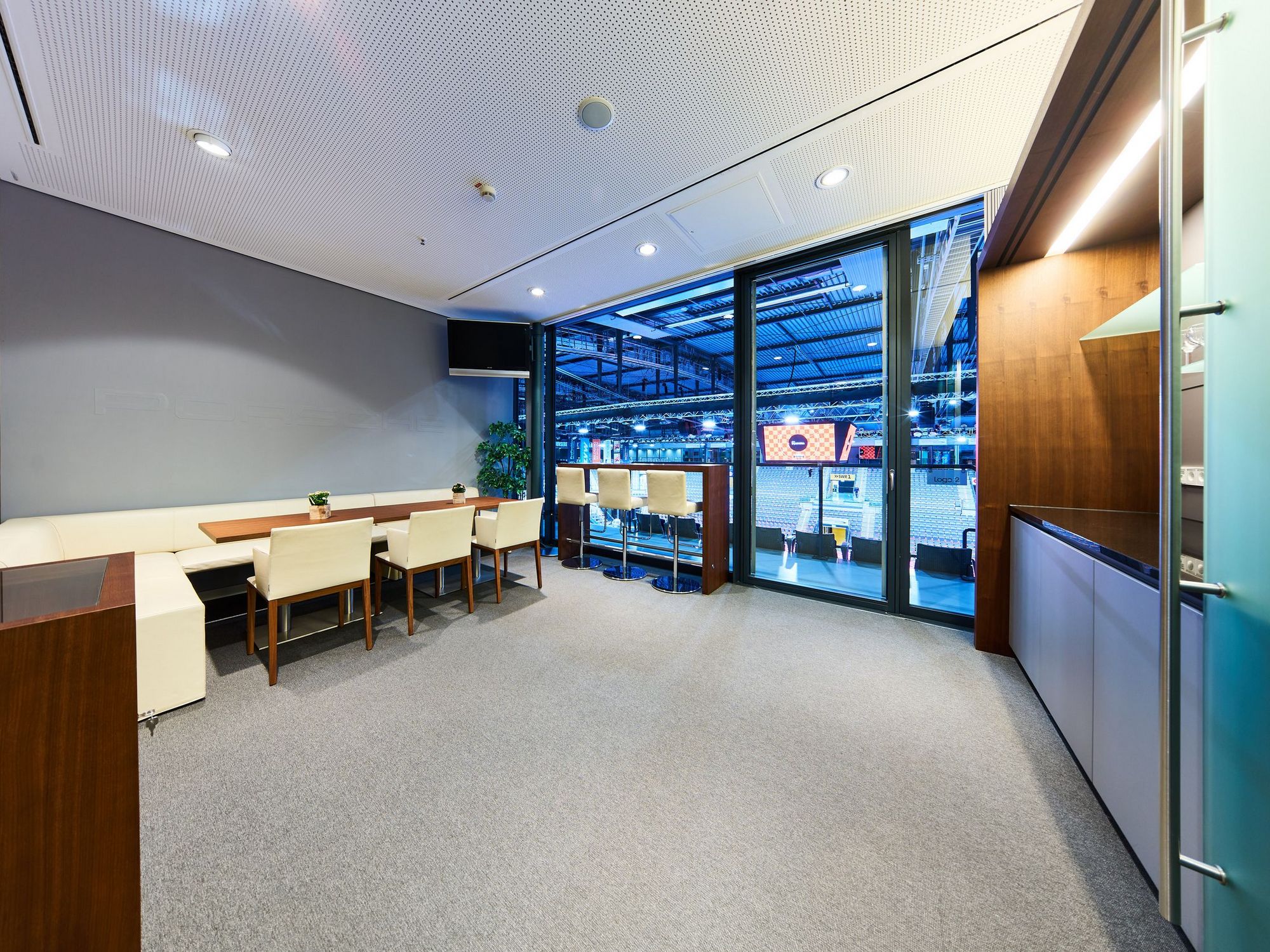 Blick in eine Loge mit einem Thresen auf der rechten Seite, links einem kleinen Kühlschrank und weißen Stühlen an einem langen Tisch. Große Fenster lassen direkt in die Porsche-Arena blicken.