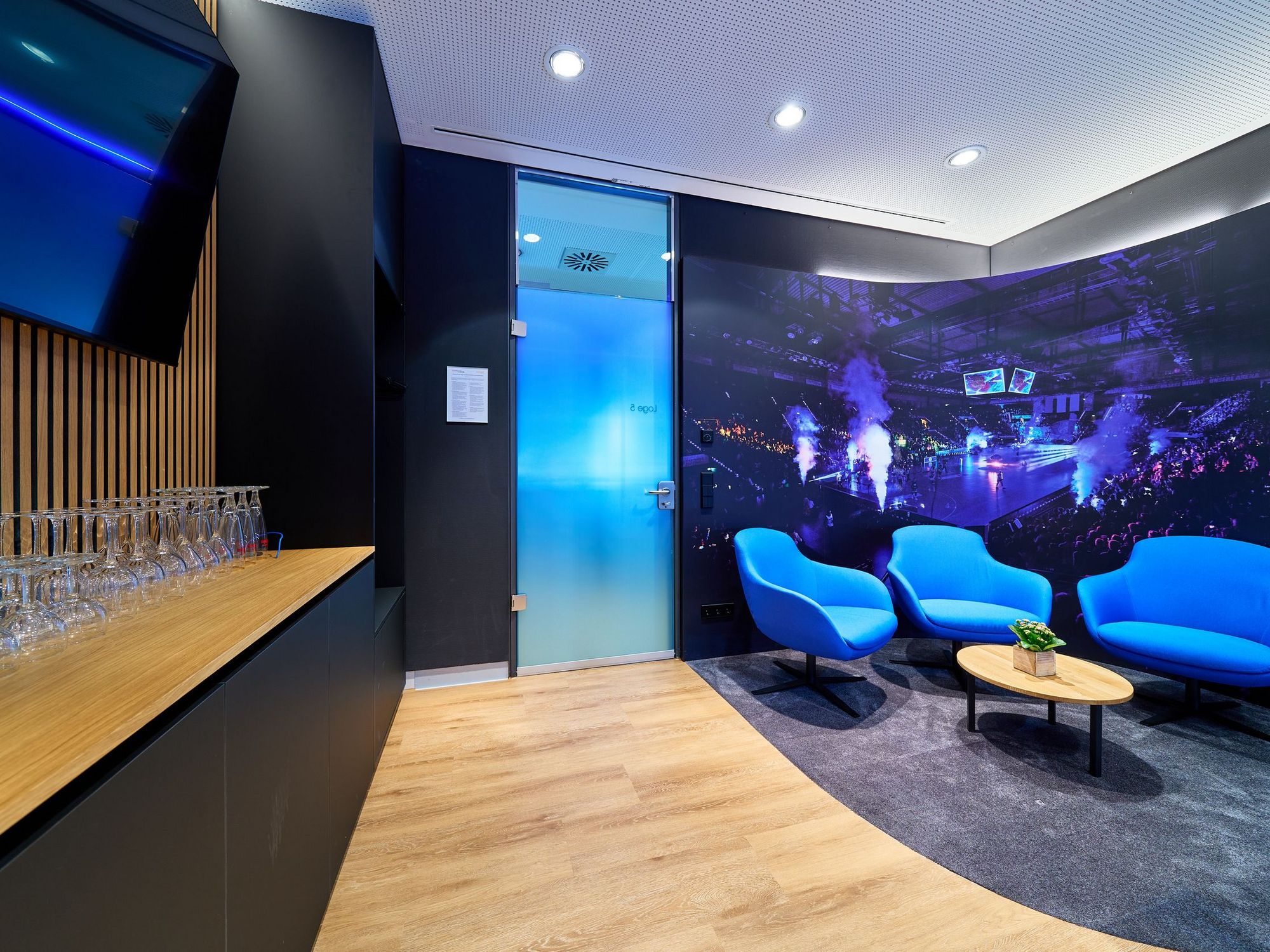 Links steht ein Thresen mit Gläsern, darüber hängt ein Fernseher an der Wand. Rechts stehen blaue Sitzmöbel um einen kleinen Tisch.