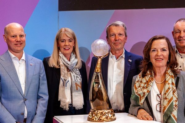 Vertreter und Vertreterinnen der Austragungsstätten stehen um den Pokal herum und lachen in die Kamera.