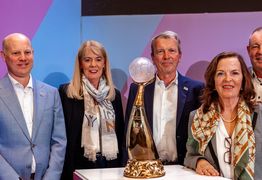 Vertreter und Vertreterinnen der Austragungsstätten stehen um den Pokal herum und lachen in die Kamera.