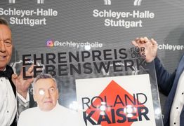 Roland Kaiser und René Otterbein halten den Ehrenpreis in Form einer Plexiglasplatte und lachen in die Kamera.